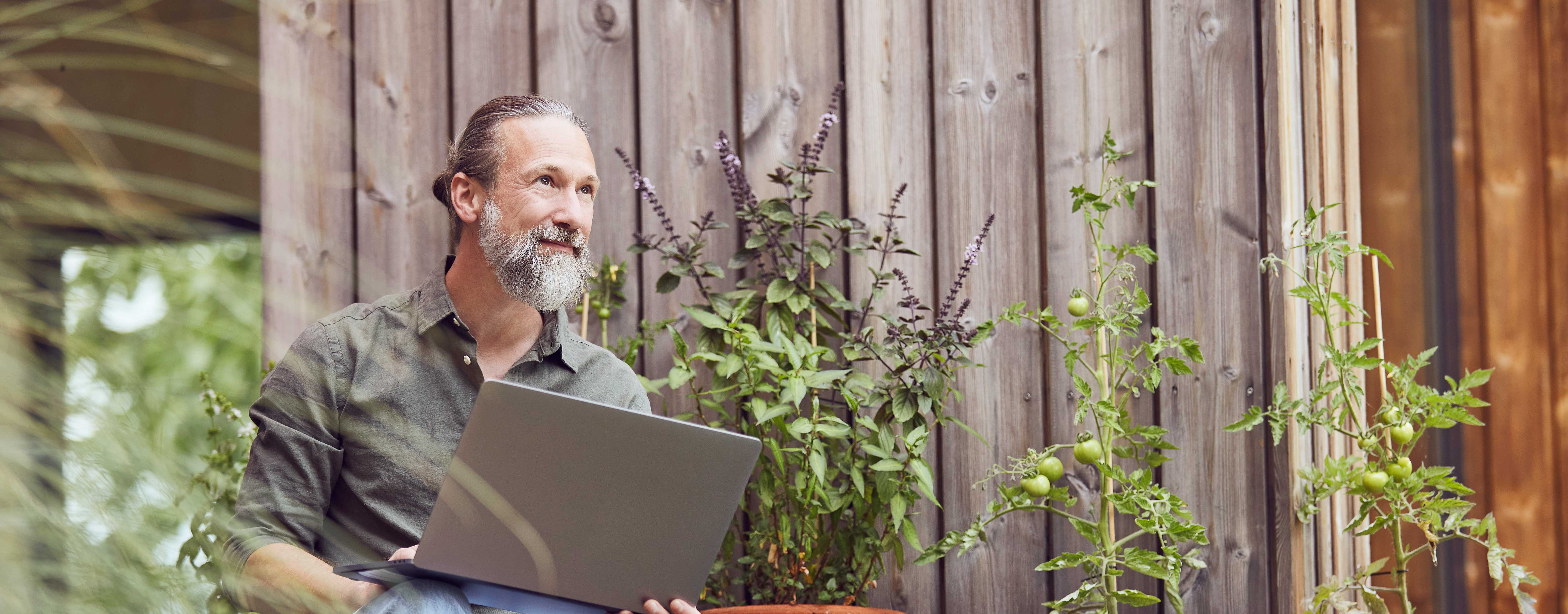 Ein Mann sitzt mit seinem Laptop vor einem Haus, inmitten von verschiedenen Pflanzen