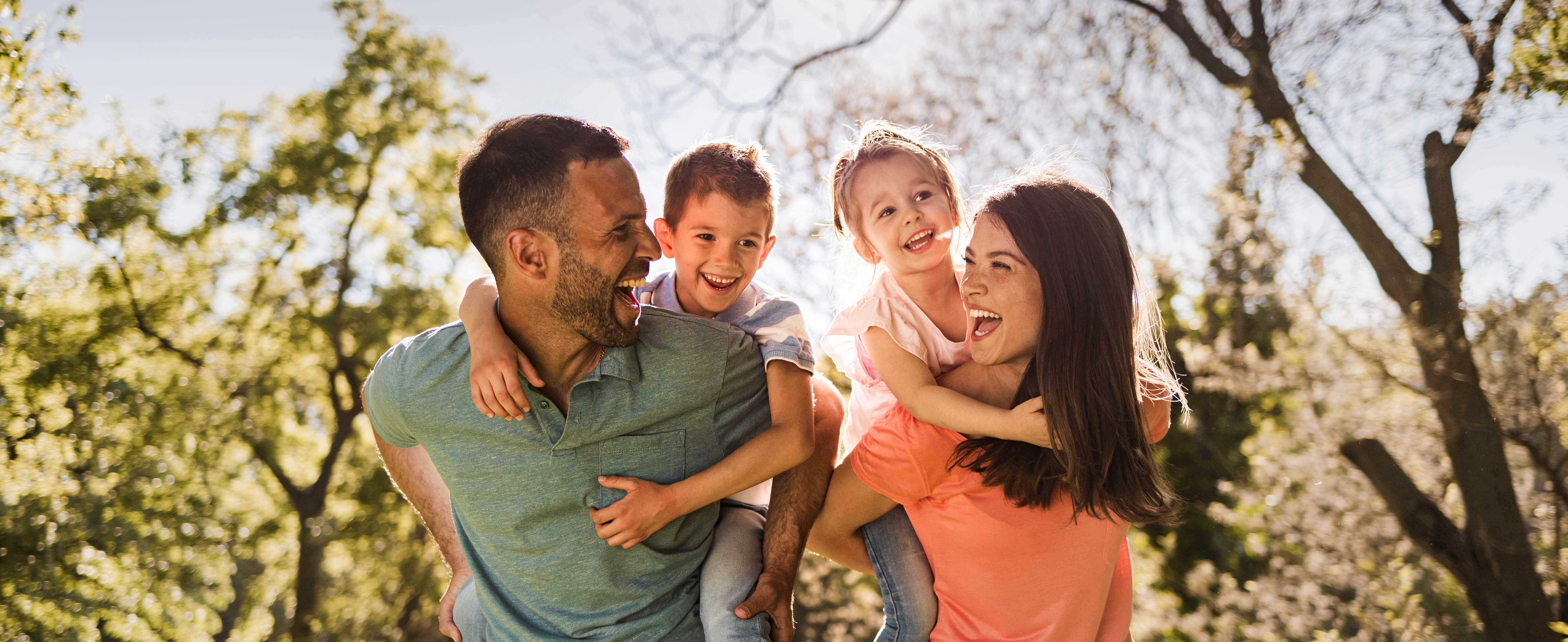 Eltern tragen ihre Kinder Huckepack und lachen sich an, im Hintergrund sind Bäume
