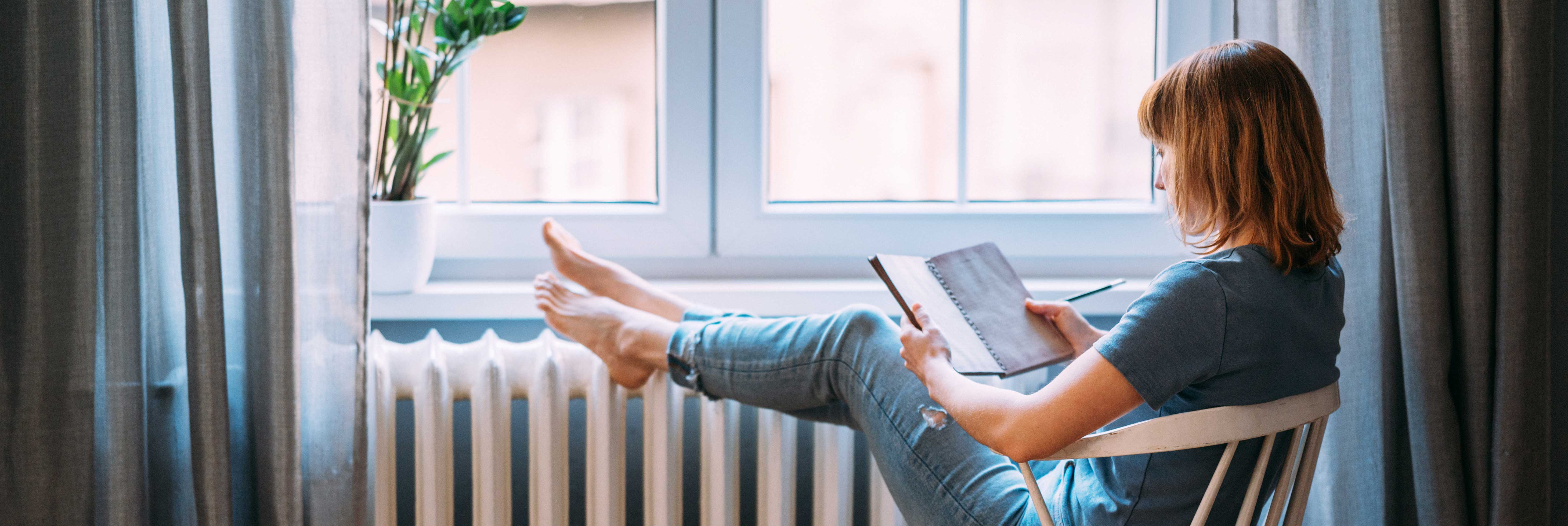 Eine Frau sitzt mit einem Notizbuch in der Hand auf einem Stuhl am Fenster und legt ihre Füße auf der Heizung ab